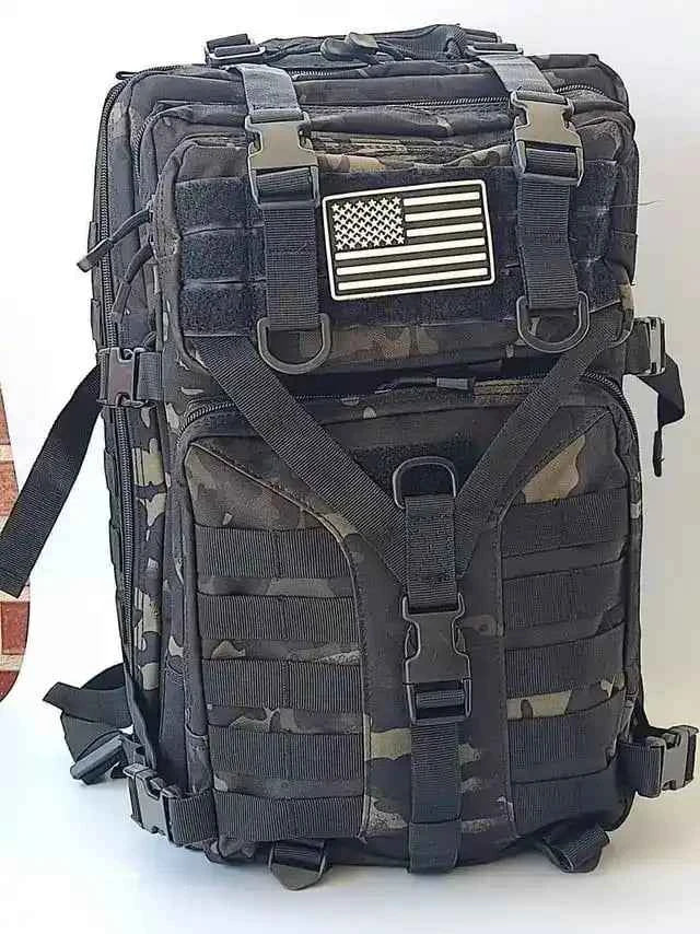 Mochila táctica camuflada negra con asa superior y parche de bandera frontal.